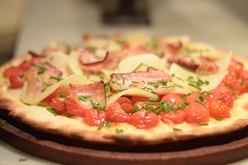 La versión de otoño de El Almacén de Pizzas. Un primer plano donde se aprecian todos sus ingredientes. Tomatitos enteros asados, láminas de papa, panceta y queso.