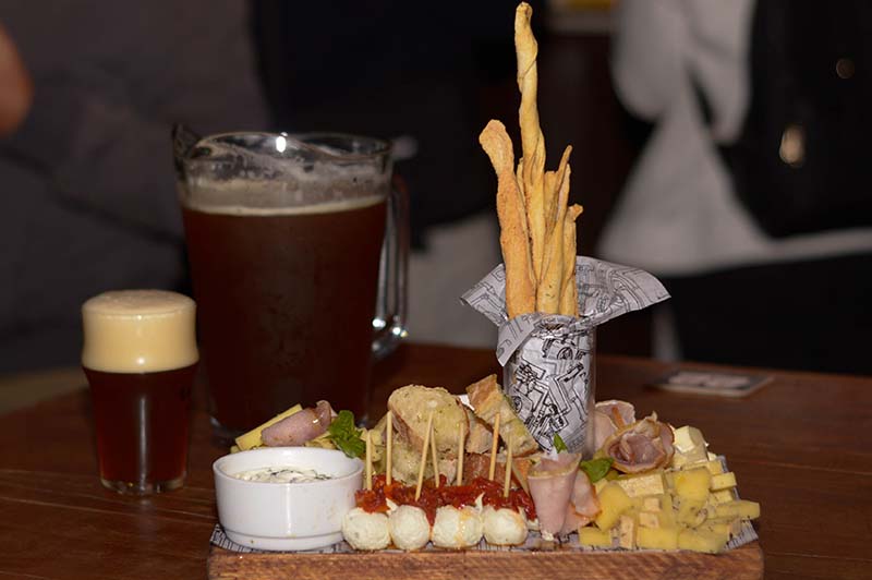 La cerveza Mensualero, jarra y vaso, aparecen junto a una nutrida tabla de quesos variados, jamones, pan ciabatta y palitos.