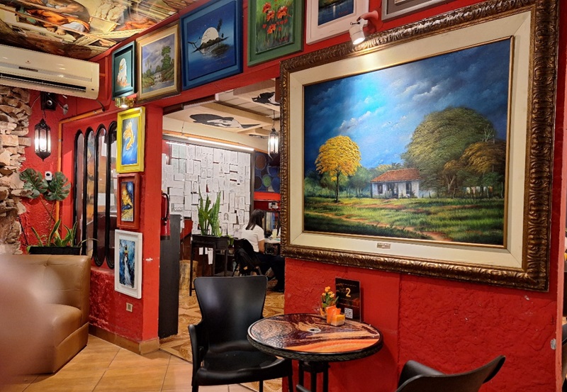 Obras con diversas temáticas cuelgan de las paredes. En el fondo se divisa el mural donde los clientes cuelgan los dibujos que realizan mientras consumen en la cafetería.